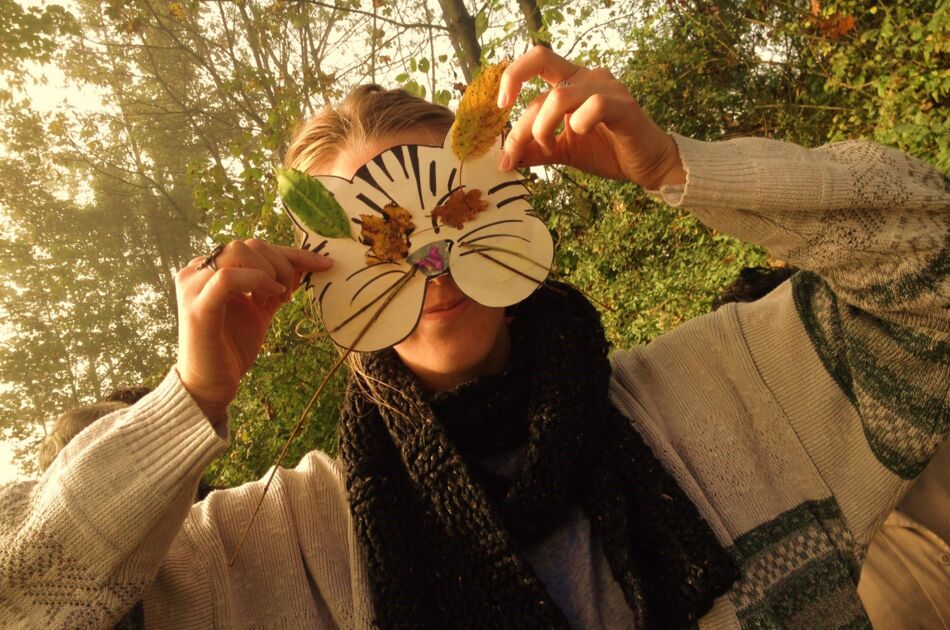 Eine Person hält mit beiden Armen eine Katzenmaske sich vors Gesicht, verziert mit Herbstblättern